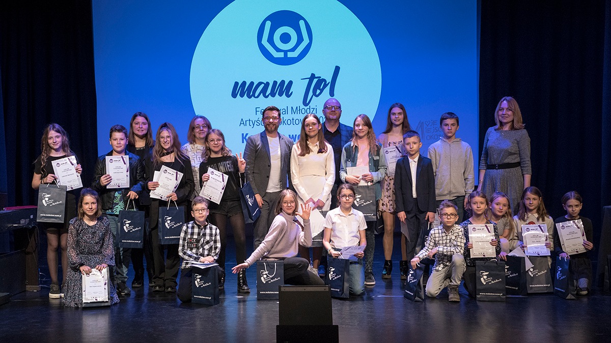 Na zdjęciu: grupa osób na scenie na tle ekranu z logo Konkursu Wokalnego MAM to! 2022; z przodu kuzające dzieci z dyplomami laureatów, za nimi stojący jurorzy i inne dzieci z dyplomami.