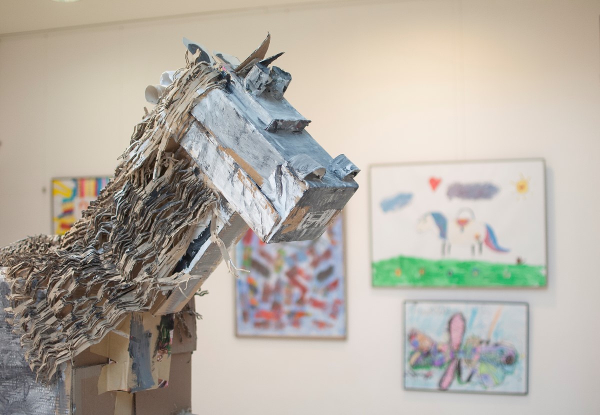 Zdjęcie pracy wykonanej na warsztatach plastycznych. Na zdjęciu znajduje się koń wykonany z kartonów. Widoczna jest jedynie glowa oraz szyja konia w lewej części kadru. W tle widoczne prace plastyczne oprawione w antyramy, wiszące na białej ścianie.