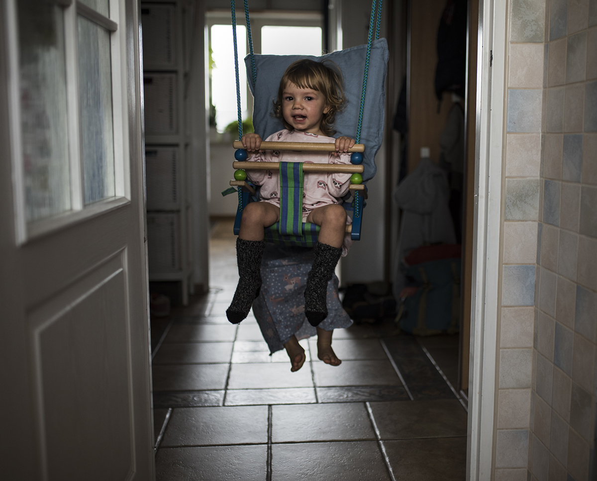 Rama w fotografii: dziecko buja się na huśtawce domowej zawieszonej pomiędzy futrynami, jest jednocześnie na tle prostokąta utworzonego z rozjaśnionego okna. Fot. Zuzanna Jach