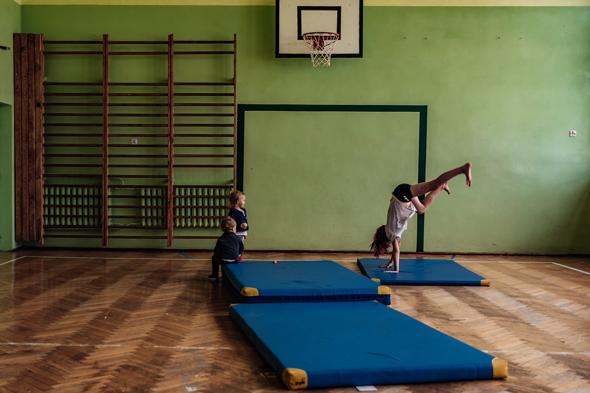 Geometryczna rama w fotografii: nastolatka w sali gimnastycznej robi gwiazdę na tle prostokąta w roli bramki wymalowanego na ścianie, przyglądają jej się dwa maluchy. Fot. Agnieszka Mocarska
