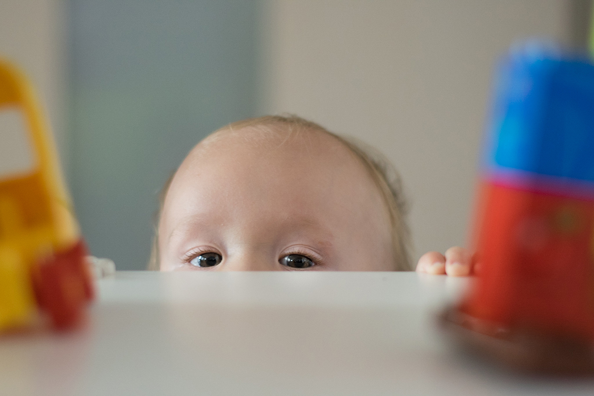 Rama zbudowana perspektywą: spomiędzy kolorowych zabawek na stole spogląda w obiektyw para dziecięcych oczu, reszta twarzy jest schowana pod stołem. Fot. Zuzanna Jach