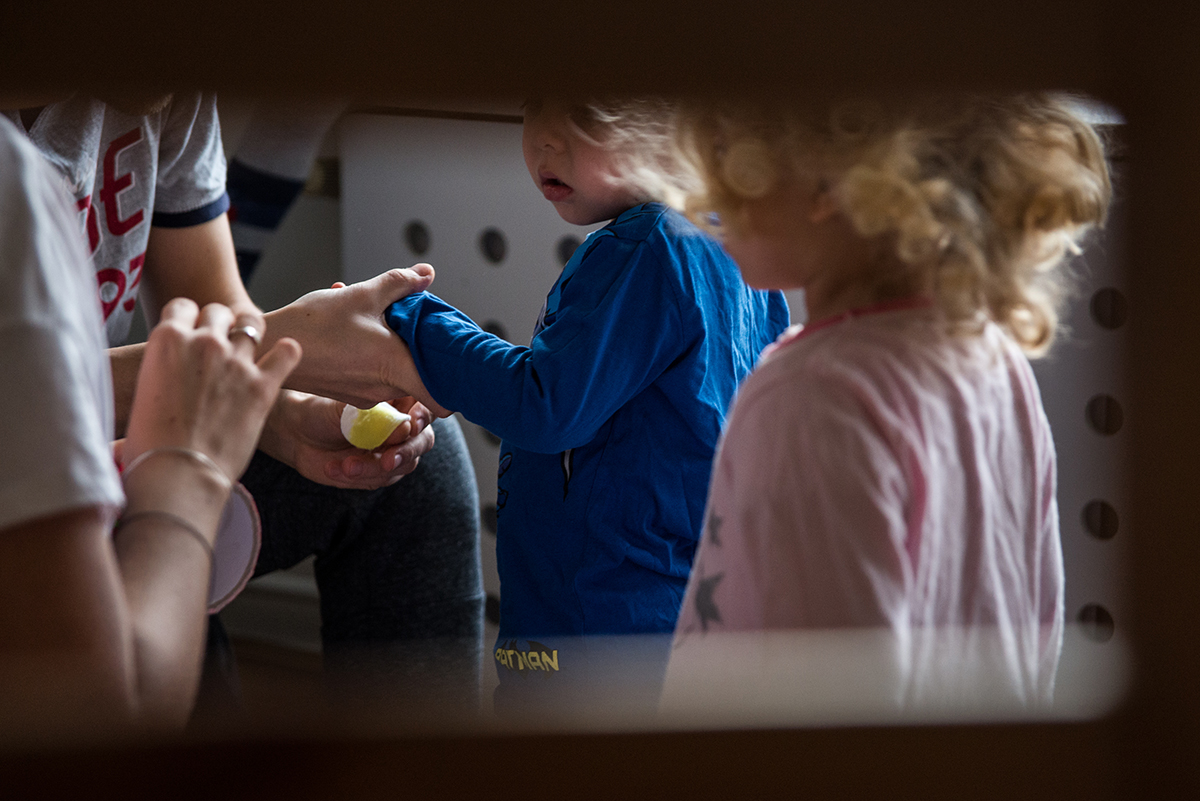 Rama dająca efekt podglądania: spomiędzy szczebelków krzesła widać dłonie rodziców naklejających plasterek maluchowi, obok niego stoi jego siostra. Fot. Agnieszka Mocarska