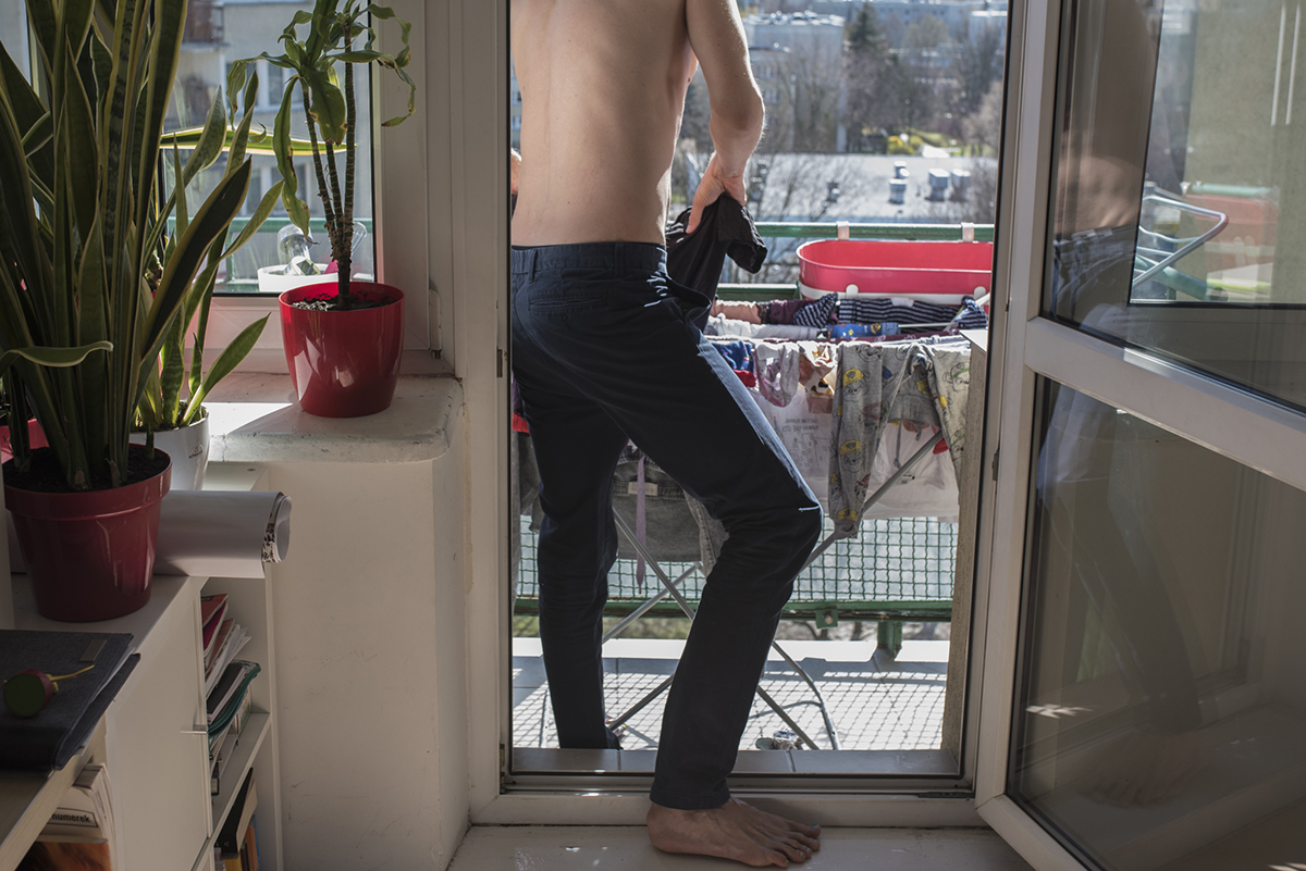 Rama dodająca kontekstu środowiskowego: w drzwiach balkonowych stoi mężczyzna i wiesza na suszarce wstawionej na balkon ubranie, w oddali widać osiedle. Fot. Zuzanna Jach