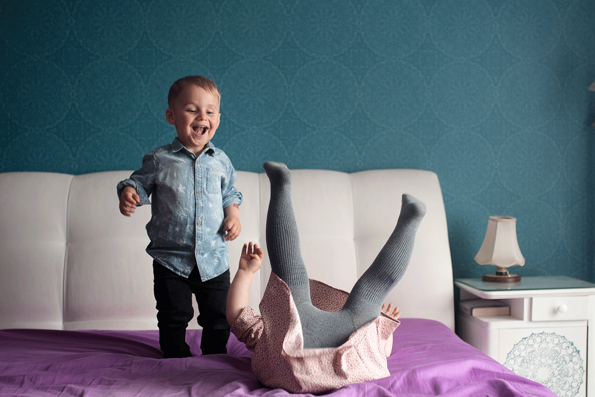 Na zdjęciu widać 2-letniego śmiejącego się chłopca po lewej, po prawej widać 4-letnią dziewczynkę robiącą fikołka na łóżku. fot. Zuzanna Jach