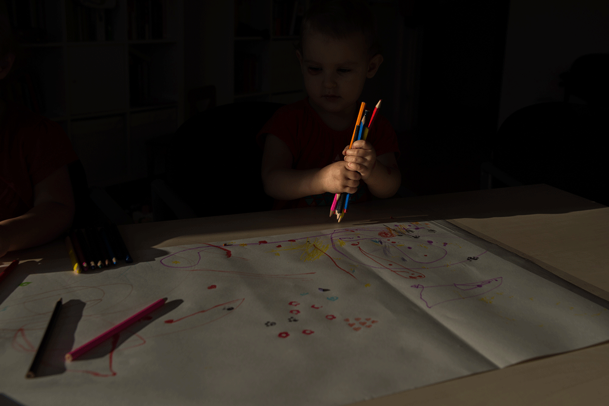 Na zdjęciu widnieje duża karta, a na niej rysunki. Po lewej stronie kadru znajduje się 2-letnia dziewczynka trzymająca pęk kredek. Jej twarz jest w cieniu. Fot. Agnieszka Mocarska