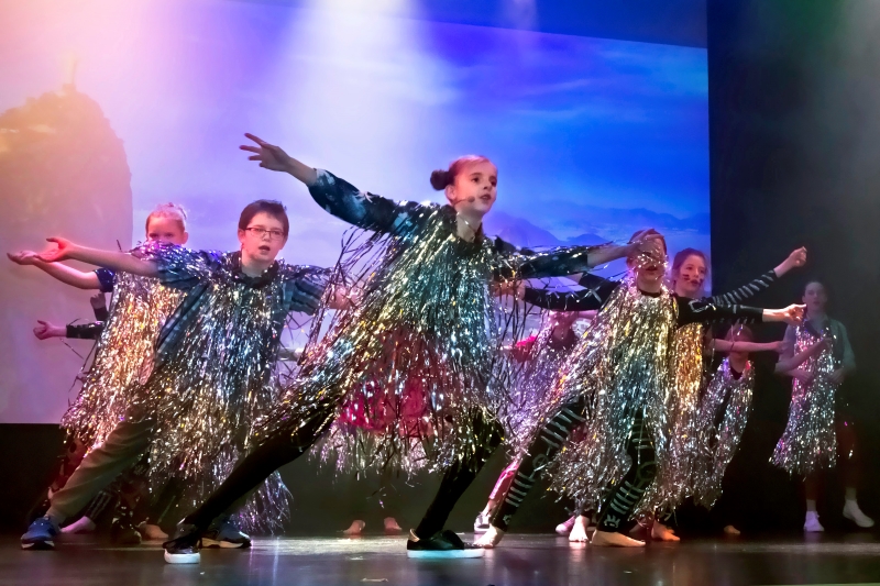 Na zdjęciu grupa dzieci występujących na scenie. Ubrani w kolorowe, błyszczące stroje. Dzieci stoją równo z pozie tanecznej.   fot. W. Hansen