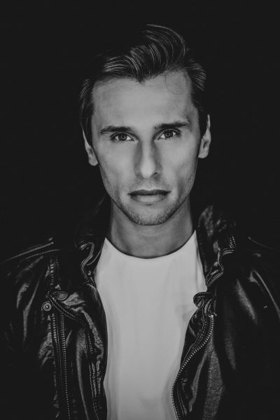 Czarno-biały portret mężczyzny. Czarne włosy, ubrany w białą koszulke oraz czarną kurtkę skórzaną. Patrzy prosto w obiektyw. fot. Ewelina Petryka