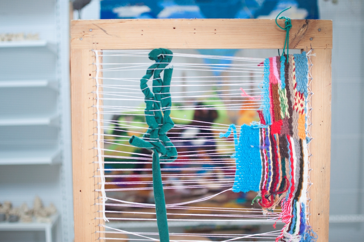 Zdjęcie pracy uczestnika zajęć. Na zdjęciu w drewnianej, roboczej ramie znajduje się wyplatanka ze sznurków. Różnokolorowe pasy, abstrakcyjne geometryczne formy. fot.D. Dzieniszewska