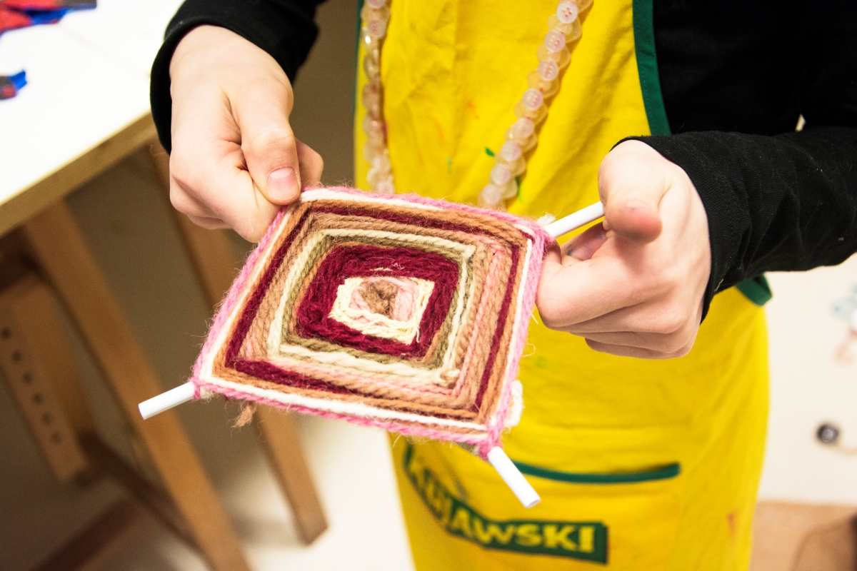 Zdjęcie pracy uczestnika. Na zdjęciu praca trzymana w rękach młodej osoby. Praca w kształcie rombu, składająca się z kolorowych sznurków. Wyplatane na drewnianych patykach.  fot. D.Dzieniszewska