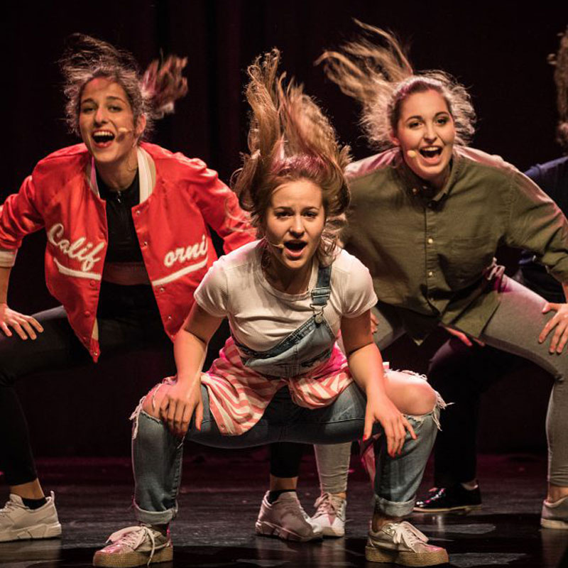 Na zdjęciu trzy dziewczyny na scenie w dynamicznych pozach. Ubrane luźno, włosy spięte. Stoją na scenie z założonymi mikrofonami nagłownymi. 