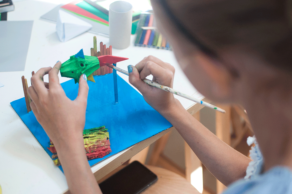 Na zdjęciu widoczne ręce młodej uczestniczki warsztatów plastycznych oraz wykonywana przez nią praca - kolorowa ryba pomalowana farbami. Praca trzymana jest nad kartką papieru pomalowaną na niebiesko jako morze. fot. Dominika Dzieniszewska