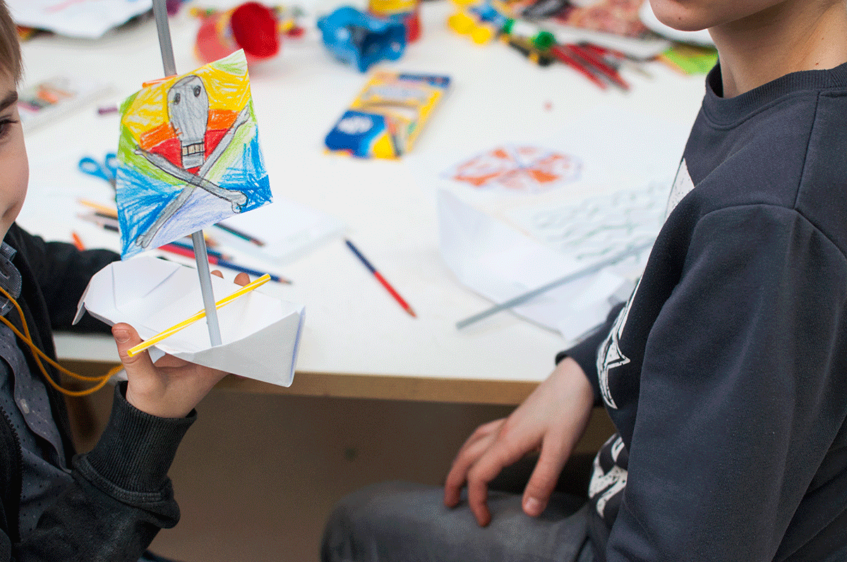 Na zdjęciu widoczny fragmenty twarzy dzieci oraz, w centralnym punkcie kadru, praca wykonana przez jednego z nich. Jest to piracki statek, wykonany z papiery i pomalowany kredkami. Na żaglu piracka czaszka. W tle biały stół z przyrządami plastycznymi.