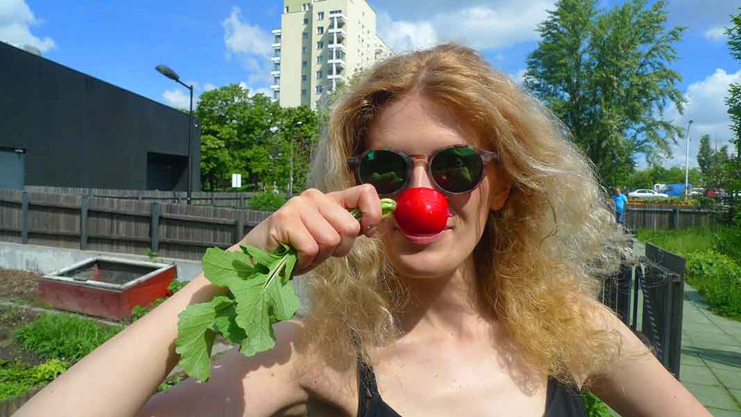 Na zdjęciu kobieta w słonecznych okularach, Klaudia, ze świeżą soczystą rzodkiewką przyłożoną do nosa. Stoi we Wspólnym Ogrodzie SDK. Za nią, w tle, fragment budynku SDK oraz osiedle Służew nad Dolinką. Fot. Dominika Dzieniszewska