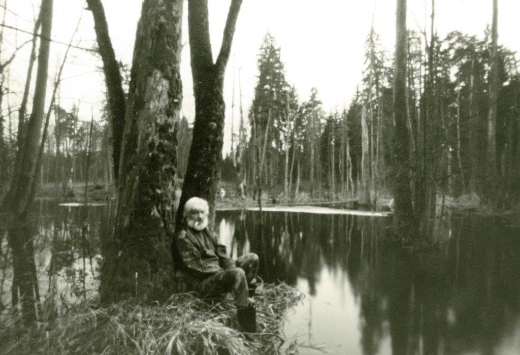 Fotografia przedstawia siedzącego na powalonym pniu Autora wystawy, na tle jego ulubionych rozlewisk bobrowych (fotografia wykonana aparatem otworkowym).