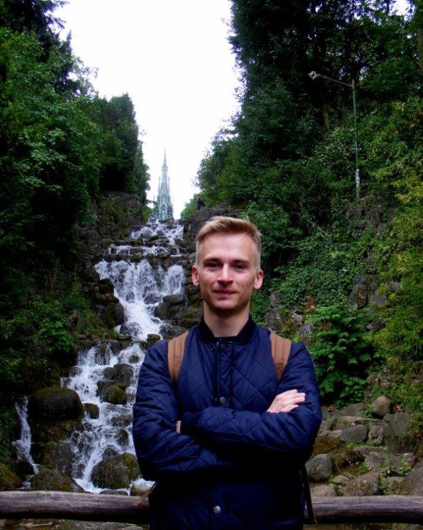 Na zdjęciu gość spotkania Łukasz Przybylak - blondyn w niebieskiej kurtce pozuje forntalnie do zdjęcia z założonymi rękami, w tle podospad skalny otoczony bujną zieloną roślinnością.