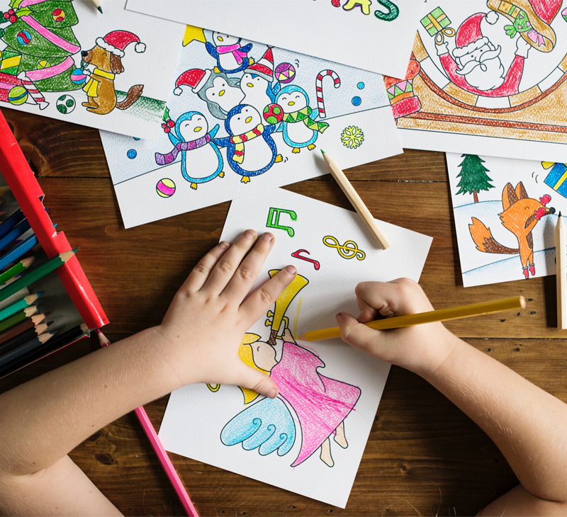 Fotografia przedstawia blat stołu, na którym widać obrazki narysowane przez dziecko. Widać również fragment rąk najprawdopodobniej dziecka.