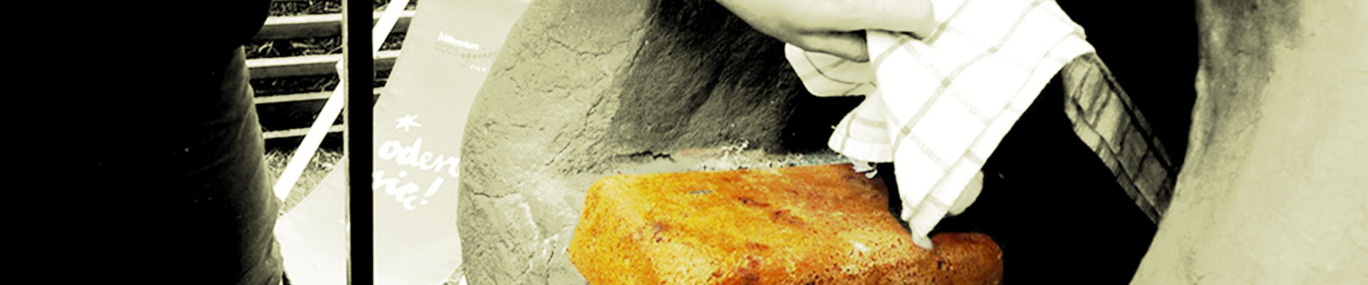 Zdjęcie: Fragment glinianego pieca chlebowego w ogrodzie SDK. Widoczne kobiece ramię, ze ściereczką w dłoni wyciągające z niego upieczony prostokątny chleb. Fot. Dominika dzieniszewska