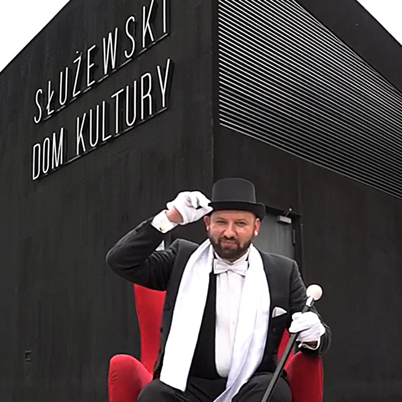 Na tle fasady SDK czerwony fotel i elegancki mężczyzna we fraku, cylindrze białym szaliku i rękawiczkach.