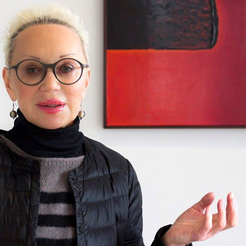 Na obrazku zdjęcie portretowe Moniki Małkowskiej na tle abstrakcyjnego obrazu olejnego w galerii obrazów. Na obrazie przeważa kolor czerwony (tło) oraz czarny - kwadrat na tle.