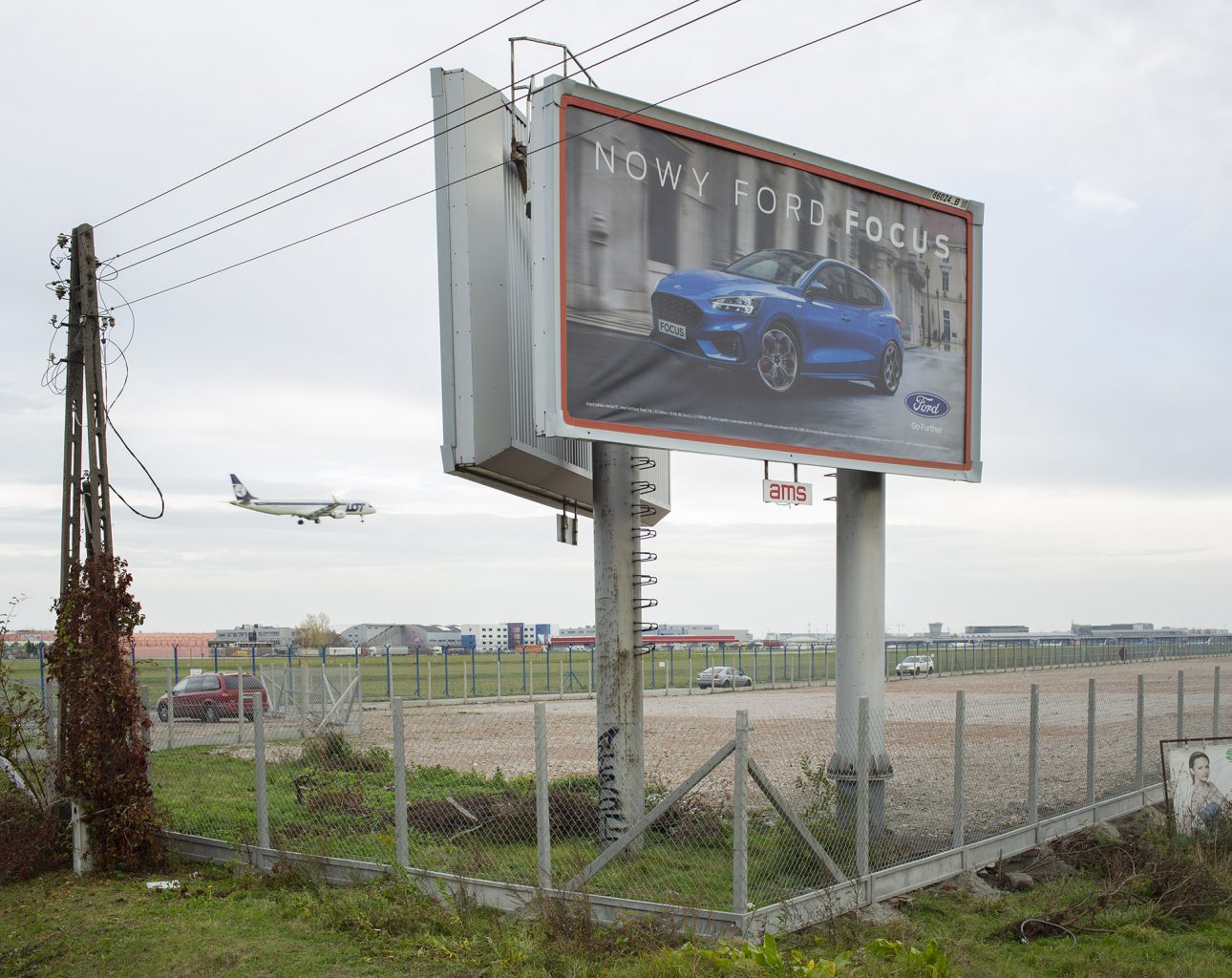 Na zdjęciu płyta lotniska widziana zza ogrodzenia z siatki. Na terenie lotniska, na pierwszym planie ogromne billboardy z reklamą samochodu osobowego. 