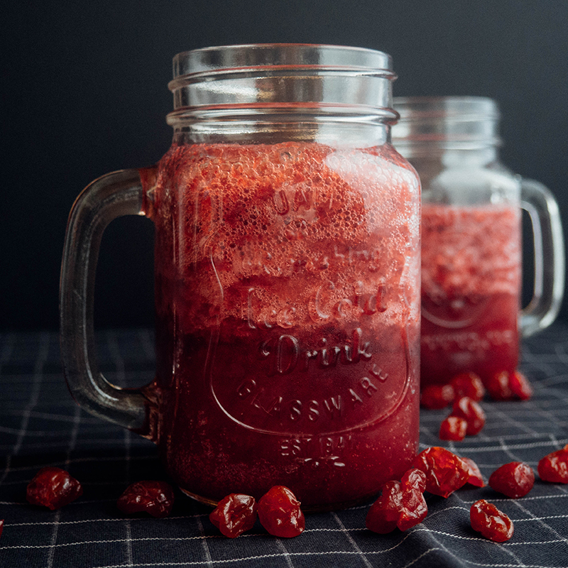 Na zdjęciu zakręcany szklany słoik z uchem, w którym znajduje się dżem z czerwonych owoców. Obok słoika porozrzucane owoce. W tle kolejny słoik.