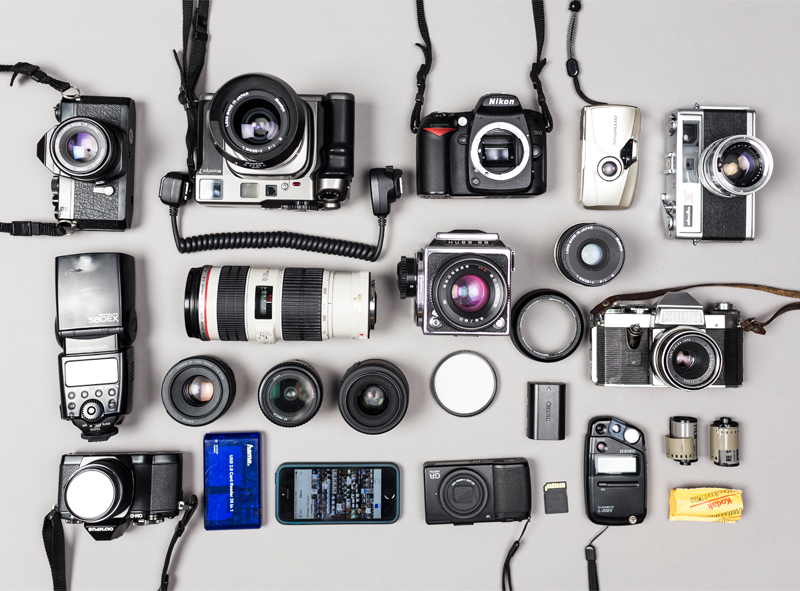 Na zdjęciu sprzęt fotograficzny rozłożony na białym blacie. Różne aparaty fotograficzne, obiektywy, lampy błyskowe, etc.