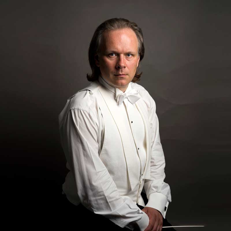 Fotografia przedstawia portret studyjny dyrygenta w stroju koncertowym. Na pierwszym planie biała koszula, biała kamizelka i biała mucha na szyi muzyka. Ręce złożone na nowach, w dłoni trzyma batutę. Włosy ułożone do tyłu. Tło szarobrunatne.