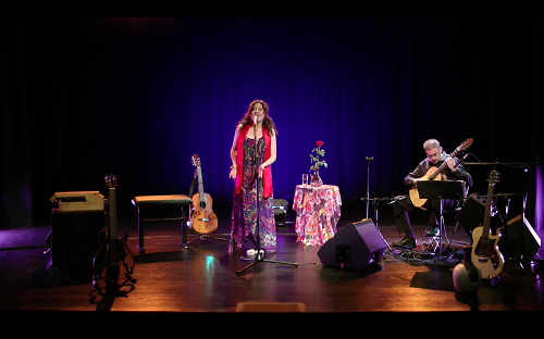 na zdjęciu Anna Riveiro i Michał Pindakiewicz w trakcie nagrania koncertu. Artystka śpiewa do mikrofonu w centralnej części zdjęcia, gitarzysta gra na intrumencie w prawym rogu. wiatło pada na piosenkarkę ubraną w długą suknię i czerowny szal. W tle instrumenty, mikrofony na statywie i granatowa kotara sceniczna.