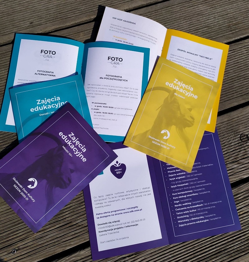 Na zdjęciu trzy rodzaje folderów na nowy sezon artystyczny. Widać sześć folderów, po dwa z każdego typu: dwa folery w kolorze ciepłej żółci dedykowany dzieciom, dwa foldery seledynowe dedykowane młodzieży i osobom dorosłym oraz dwa foldery fioletowe dedykowane seniorom. Font w ulotkach jest w kolorze ich okładek. Broszury leżą na drewnianej podłodze przy amfiteatrze SDK.