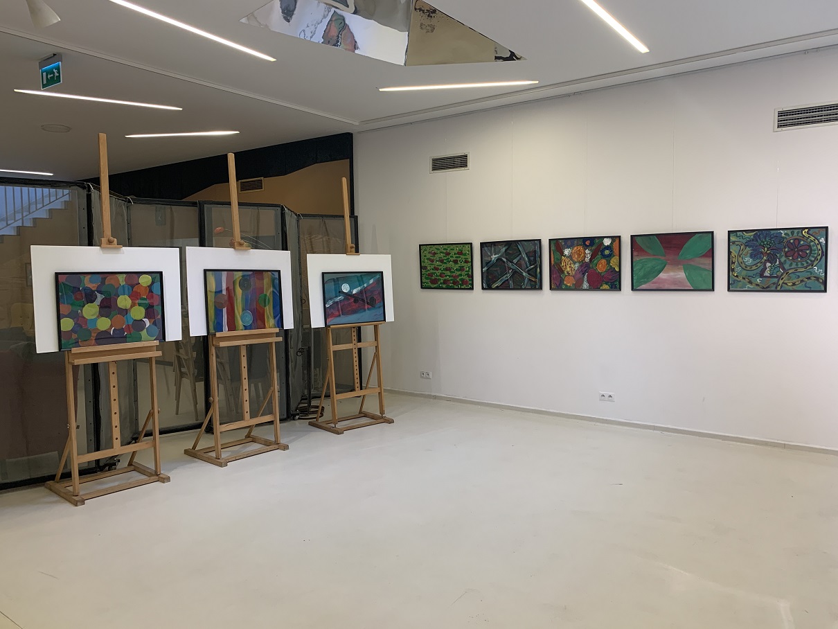 Na zdjęciu widać kilkanaście prac wywieszonych w Galerii przy Kozach, należących do uczestników projektu "Onkoprzestrzeń kreatywna". Kilka prac jest wystawionych na sztalugach, pozostałe wiszą na białej ścianie. Prace artystyczne są pokazanie w planie ogólnym, co utrudnia ich opisy. Dominującym kolorem zdjęcia jest biel.