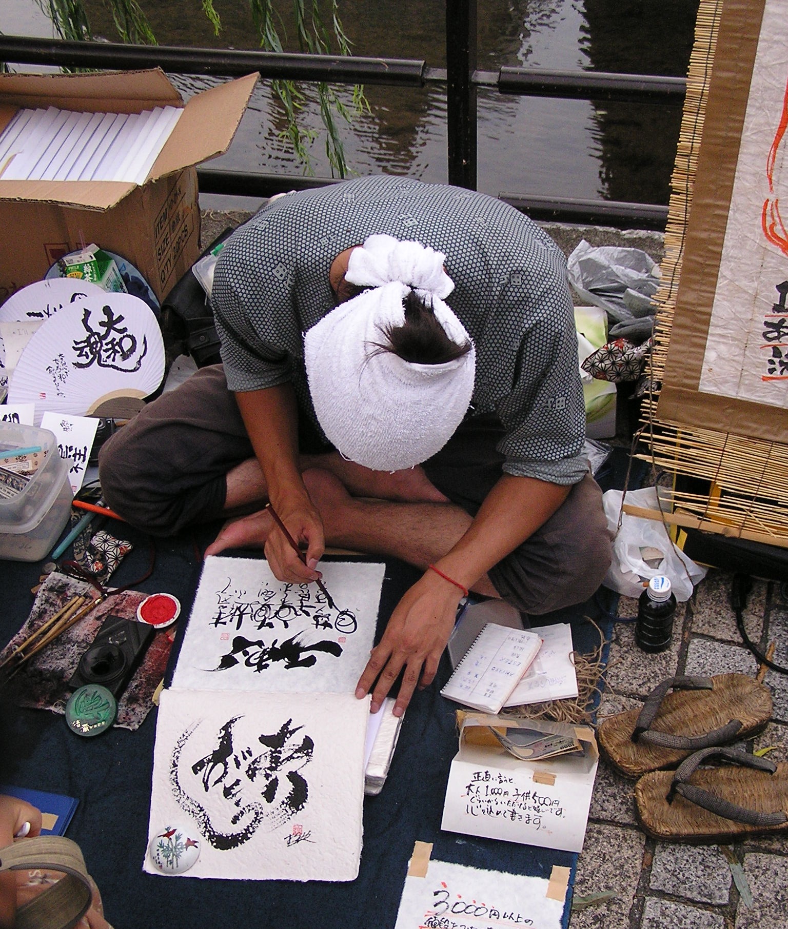 Zdjęcie przedstawia siedzącego w siadzie skrzyżnym Japończyka (ubrany w tradycyjny strój), który z pochyloną głową dokonuje ostatnich poprawek do wykonanej przez siebie tradycyjnej kaligrafii. Zdjęcie zostało wykonane na ulicach Kioto.