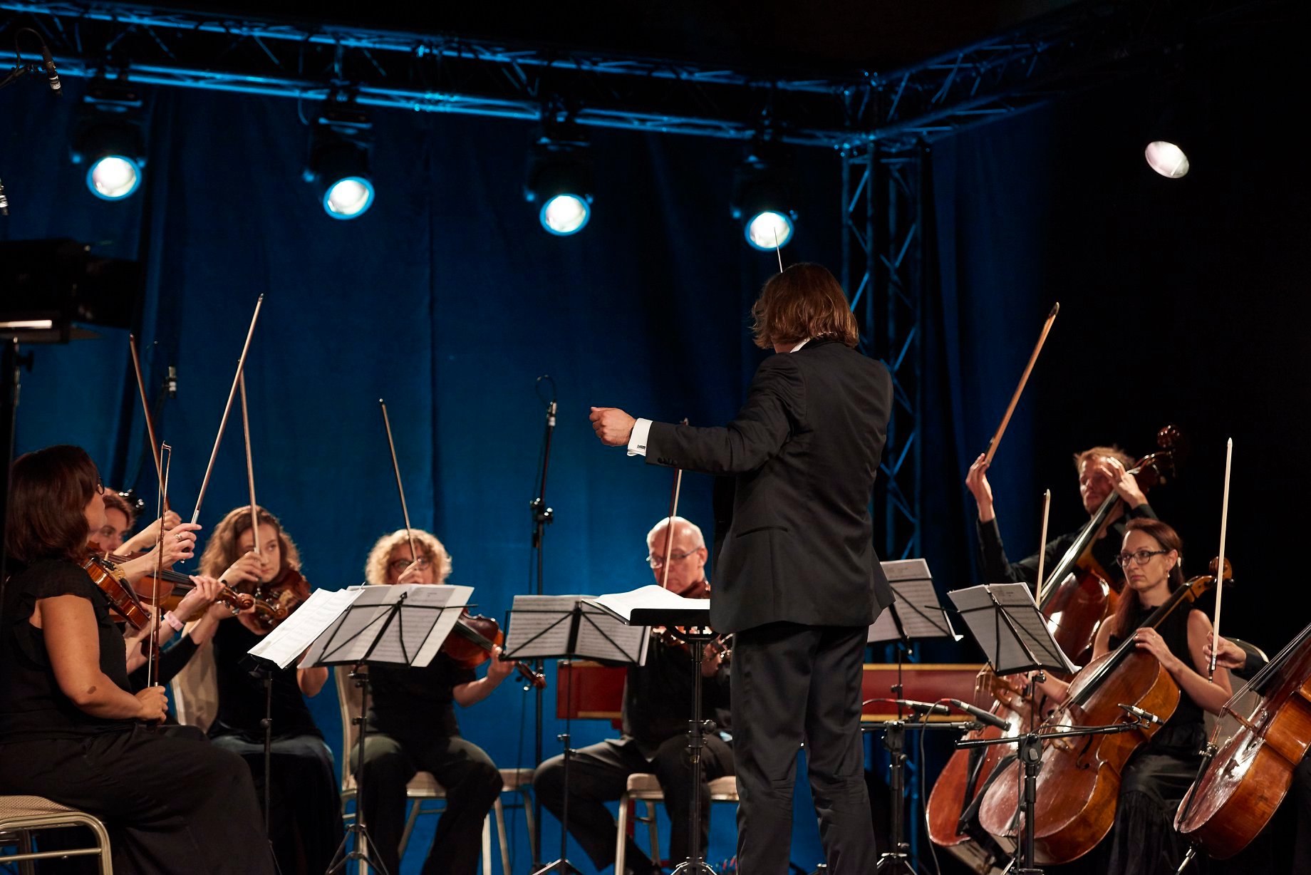grafika promocyjna Zdjęcie przedstawia dynamicznie sportretowaną orkiestrę smyczkową w czasie koncertu. Każdy trzyma przy sobie intrument, muzycy są elegancko ubrani, patrzą na rozstawione na stoikach kartki z partyturą. Dyrygent stoi nad muzykami i energicznie kieruje orkiestrą przy pomocy batuty. Twarzy nie widać. W tle widać granatowe zasłony sceniczne.