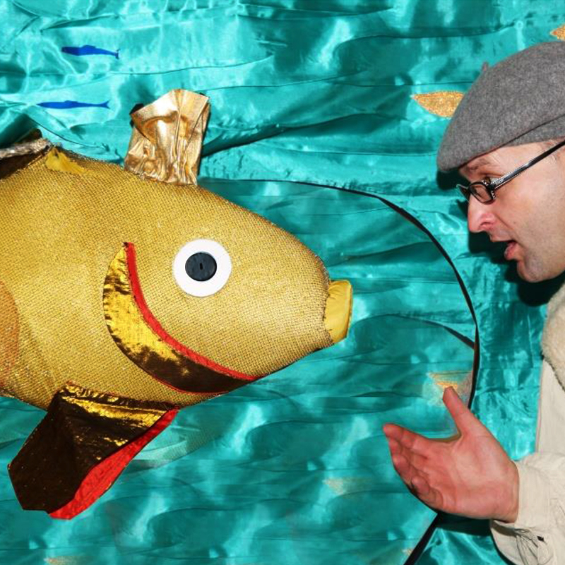 Zdjęcie przedstawia rozmawiającego aktora - odgrywającego rolę rybaka - ze złotą rybką w postaci pluszowego rekwizytu. W tle seledynowa błyszcząca tkanina imitująca wodę. 