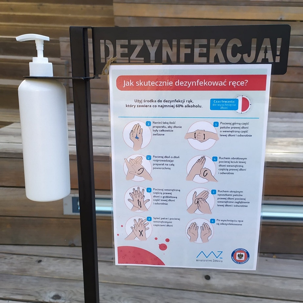 grafika promocyjna na xzdjęciu widać melatowy stojak polamowany na czarno, do którego przypięta jesy biała plastikowa butelka z dozownikiem. Stojak ma wygrawerowany napis "dezynfekcja", a do niego przypięta jest infografika Ministerstwa Zdrowia jak prawidłowo myć ręce, dołączona do artykułu. W tle drewniany amfiteatr SDK.