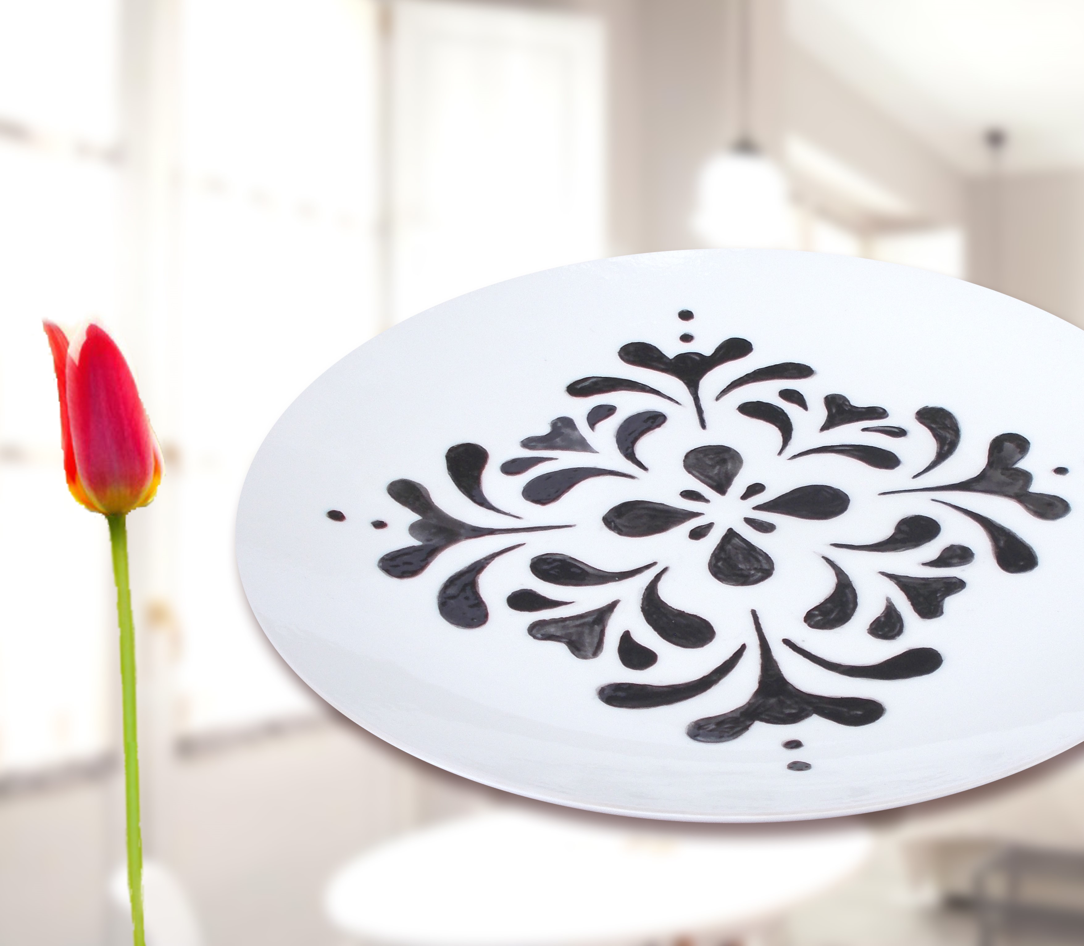Zdjęcie przestawia pracę ceramiczną Moniki Marks-Witkowskiej. Talerz jest biały, po środku widać charakterystyczne ornamenty ludowe, które rozchodzą się ku brzegom. Zdobienia są w kolorze ciemnego brązu. Po lewej stronie zdjęcia jest czerwony tulipan. W tle rozmyta przestrzeń mocno doświetlonego salonu z dominującym kolorem białym.