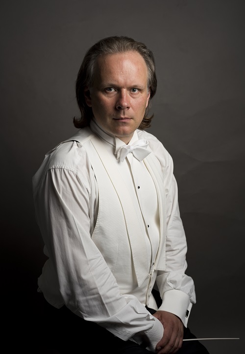 zdjęcie promocyjne Fotografia przedstawia portret studyjny dyrygenta w stroju koncertowym. Na pierwszym planie biała koszula, biała kamizelka i biała mucha na szyi muzyka. Ręce złożone na nowach, w dłoni trzyma batutę. Włosy ułożone do tyłu. Tło szarobrunatne.