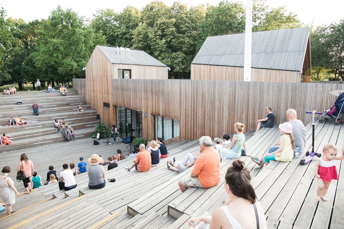 Zdjęcie przedstawia amfiteatr SDK widziany od ulicy Bacha. Po obu stronach drewnianego amfiteatru siedzi lub leży kilkanaście osób korzystających ze słonecznej pogody. W tle jasne niebo.