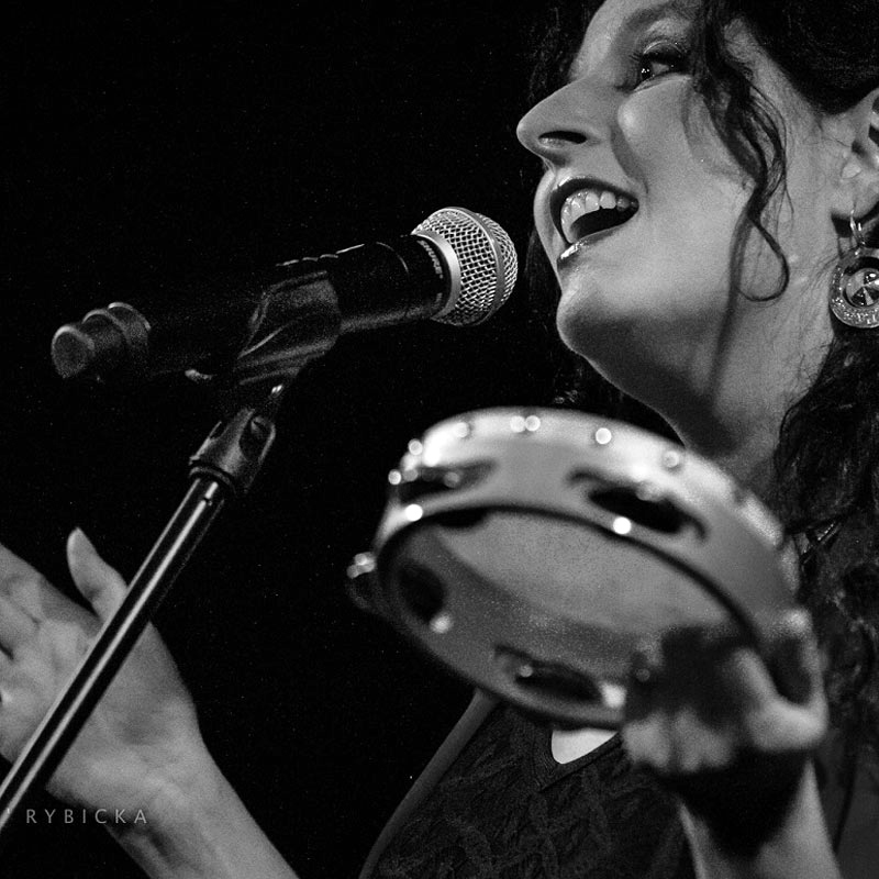Na grafice zdjęcie portretowe artystki. Twarz artystki przy mikrofonie, w ręku widoczny tamburyn. 
