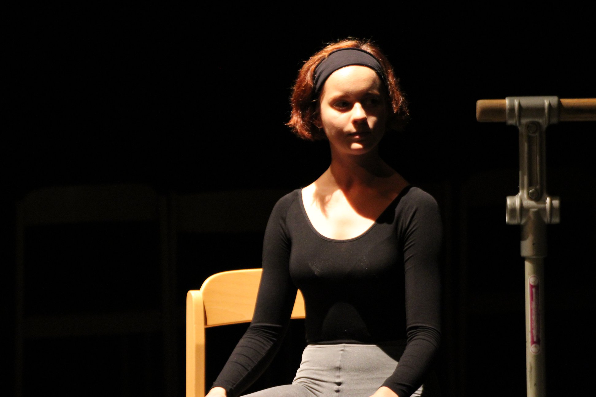 W centralnej części kadru, na krześle siedzi rudowłosa tancerka, będąca jedną z głównych bohaterek spektaklu. Ubrana jest w czarny strój do ćwiczeń. Na włosach czarna, elastyczna opaska.