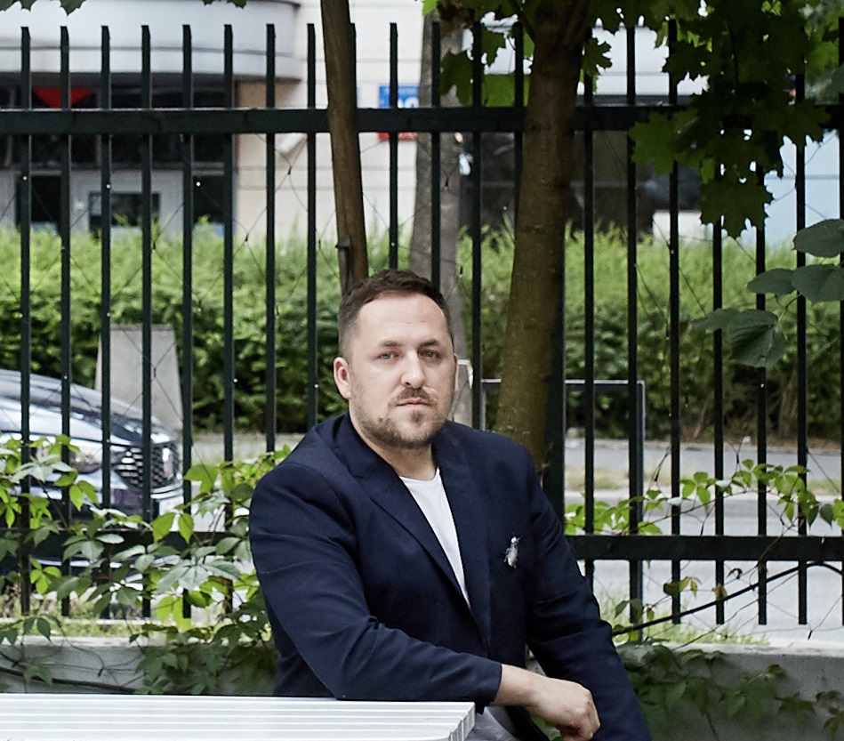 Portret gościa spotkania Na zdjęciu wizerunek gościa spotkania Pana Marcina Szczeliny. Postać siedzi za stołem, w przestrzeni miejskiej. Za nim ogrodzenie oraz fragmenty budynku oraz poruszającego się samochodu.