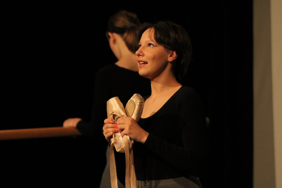 Na zdjęciu jedna z bohaterek spektaklu, trzymająca w dłoniach baletki. Za nią druga tancerka stoi tyłem, trzymając dłoń na drążku baletowym