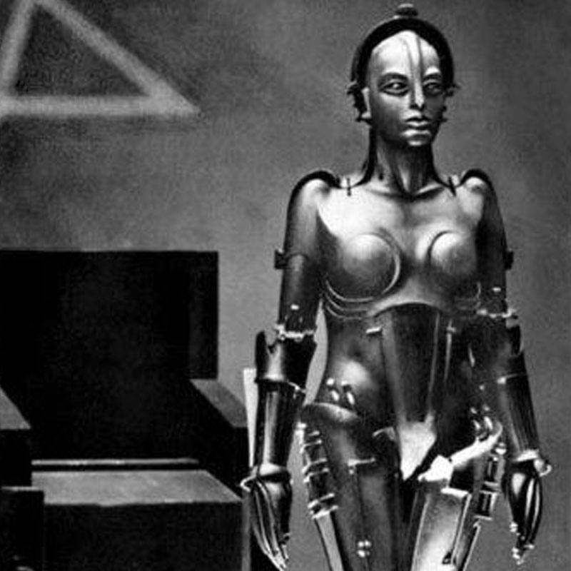 Na zdjęciu fragment pomieszczenia, w którym stoi duże krzesło oraz robot o kobiecej figurze. Całość w czerni i bieli oraz mrocznym świetle.