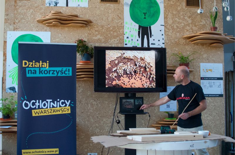 Zdjęcie przedstawia mężczyznę prowadzącego wykład na temat pszczół. W tle znajduje się roll-up programu Ochotników Warszawskich.