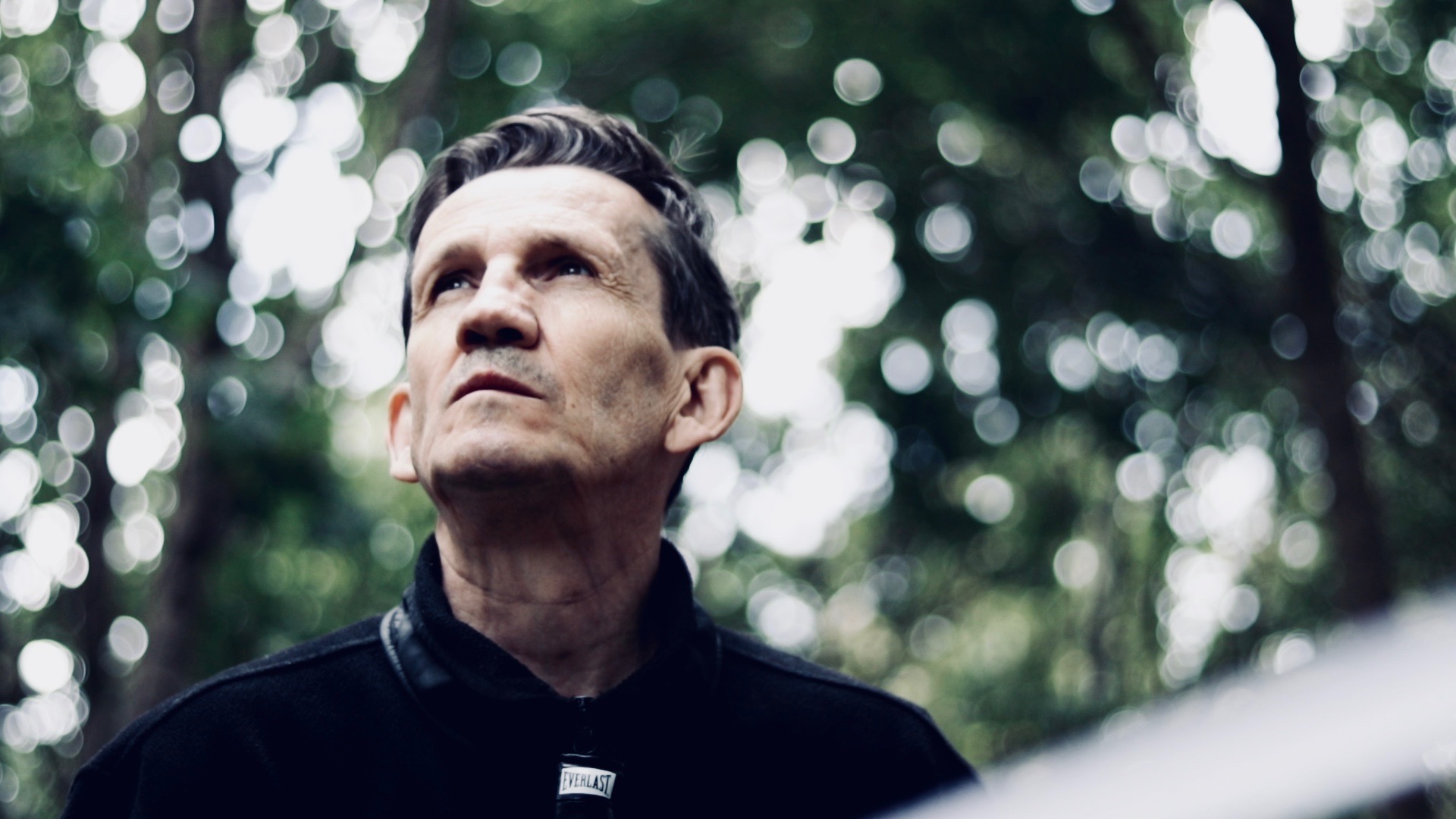 Zamyślony mężczyzna, patrzący w górę. W tle rozmyta roślinność.  