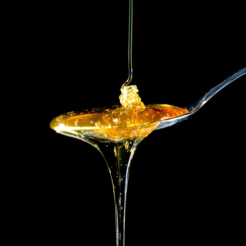 Na zdjęciu, na czarnym tle znajduje się łyżka cieknącego miodu. Miód spływa z łyżeczki. 