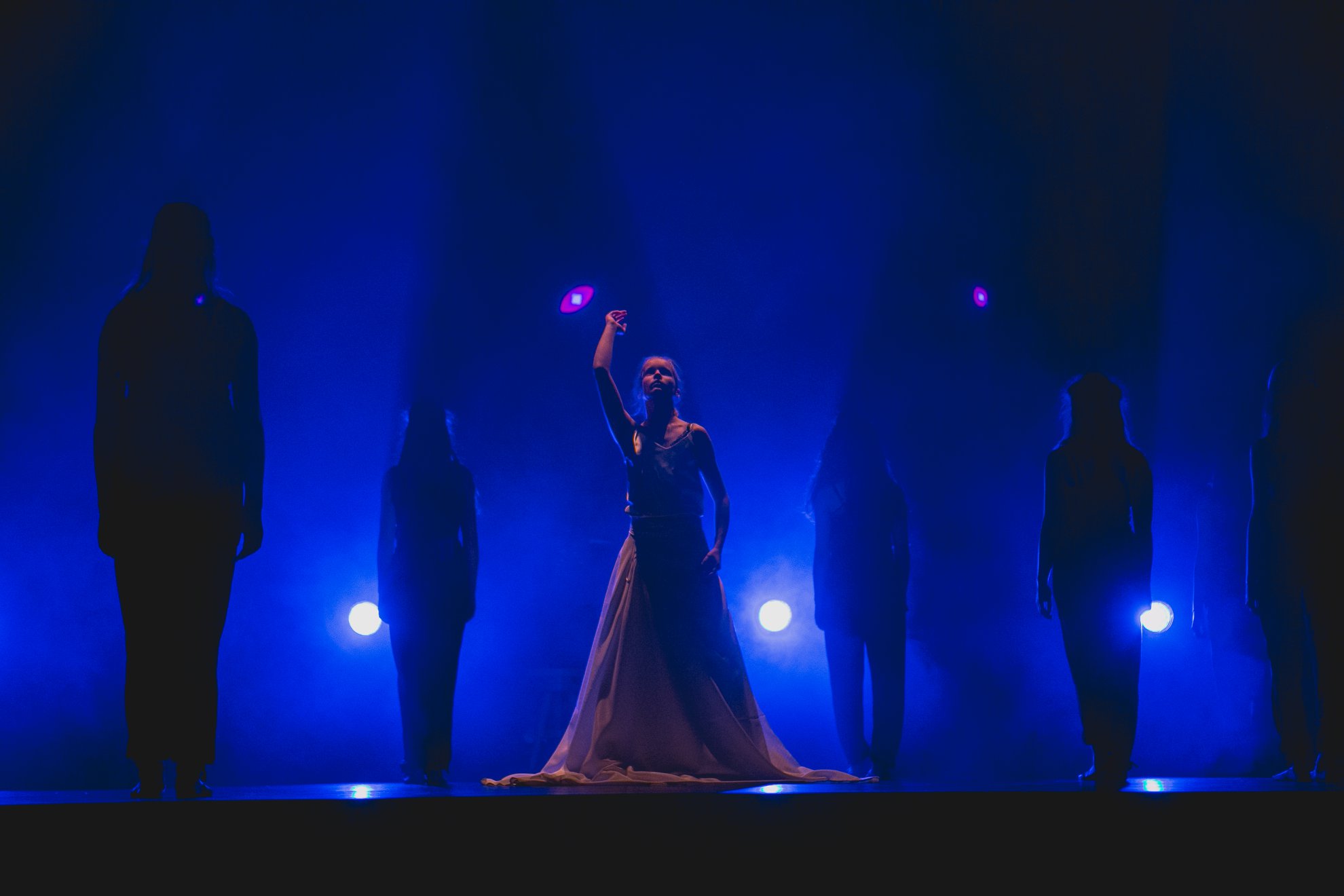 Zdjęcie promocyjne zespołu. Fot. Gorajka Foto Na zdjęciu cztery tancerki w ciemnym świetle. Pomiędzy nimi jedna tancerka w sukni, oświetlona z wyciągniętą dłonią ku górze. Tancerki znajdują się na scenie, w niebieskim świetle.