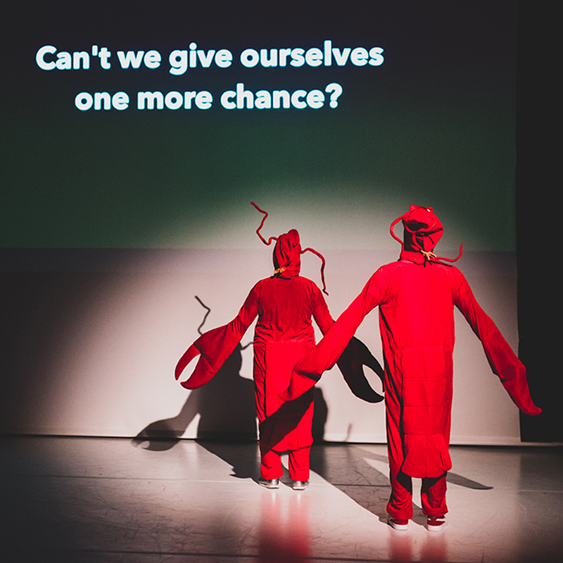 Na zdjęciu dwie postaci ubrane w czerwone skafandry, w tle ekranu biały napis w języku angielskim "Czy nie możemy dać sobie jeszcze jednej szansy?"