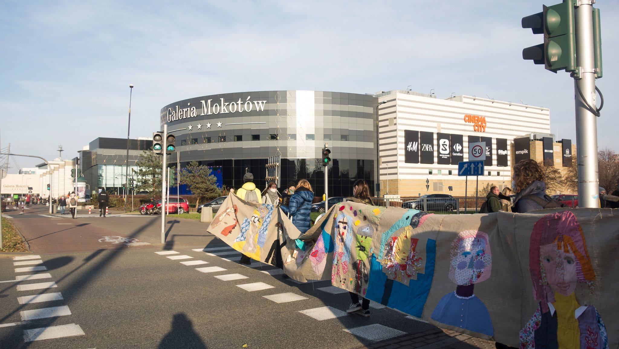 grafika promocyjna Przemarsz instalacji autoportretów wolontariuszy przez Mokotów. Uczestnicy happeningu przechodzą przez przejście dla pieszych przy Galerii Mokotów (centrum handlowe w tl)