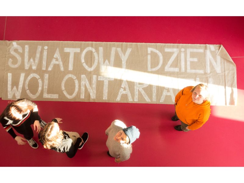 grafika promocyjna Fragment instalacji informujący o światowym dniu wolontariusza widziany od góry w Domu Kultury KADR. Zdjęcie wykonane od góry, w tle czerwona podłoga.