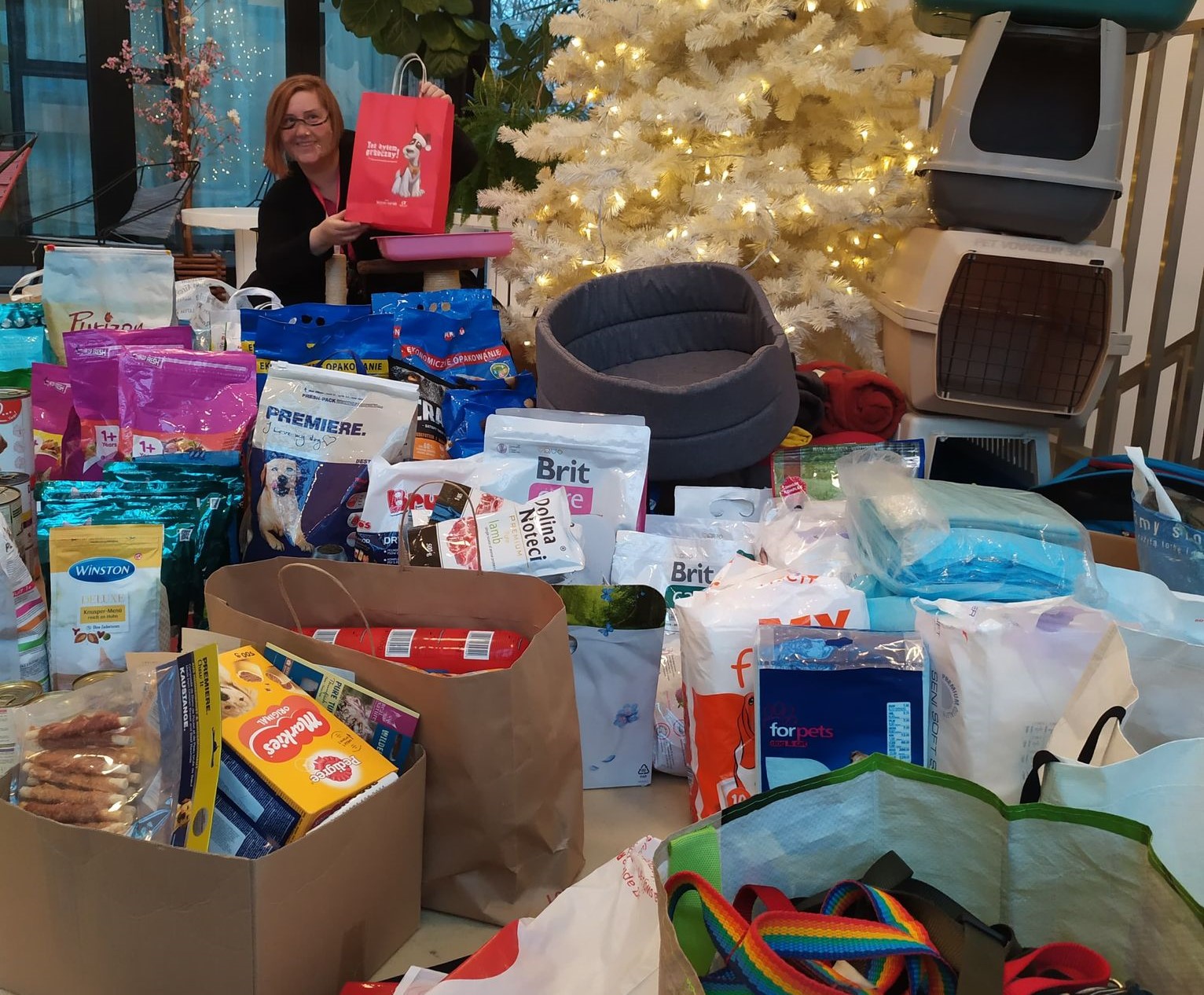 Koordynatorka zbiórki na tle przekazywanych darów. Widać kolorowe smycze,m suchą i mokrą larmę, transportery i inne. W tle choinka.